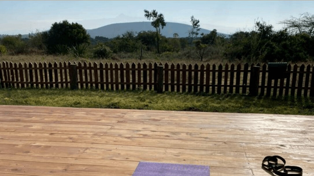 Yoga-Plattform auf der Sandai Farm mit Blick auf einen Holzzaun, grüne Landschaft und einen Berg im Hintergrund, bei klarem Morgenlicht.