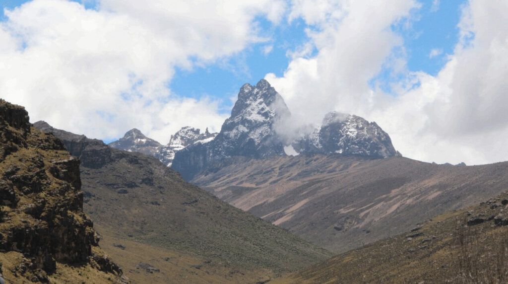 Majestätische Gipfel des Mount Kenya, bedeckt mit Schnee und umgeben von felsigen Hängen, unter einem teilweise bewölkten Himmel.