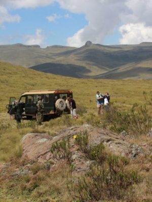 Eine Safari, organisiert von Sandai Farm African Footprints, mit einem Geländewagen und einer Gruppe von Menschen, die eine weite, hügelige Landschaft mit blauem Himmel und verstreuten Wolken erkunden.