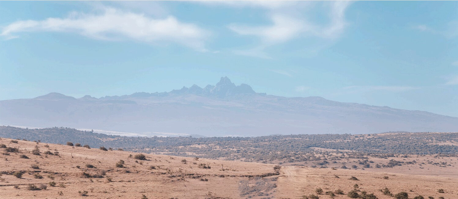 Blick auf den Mount Kenya, umgeben von trockener Savanne mit vereinzeltem Buschwerk unter einem klaren, blauen Himmel.